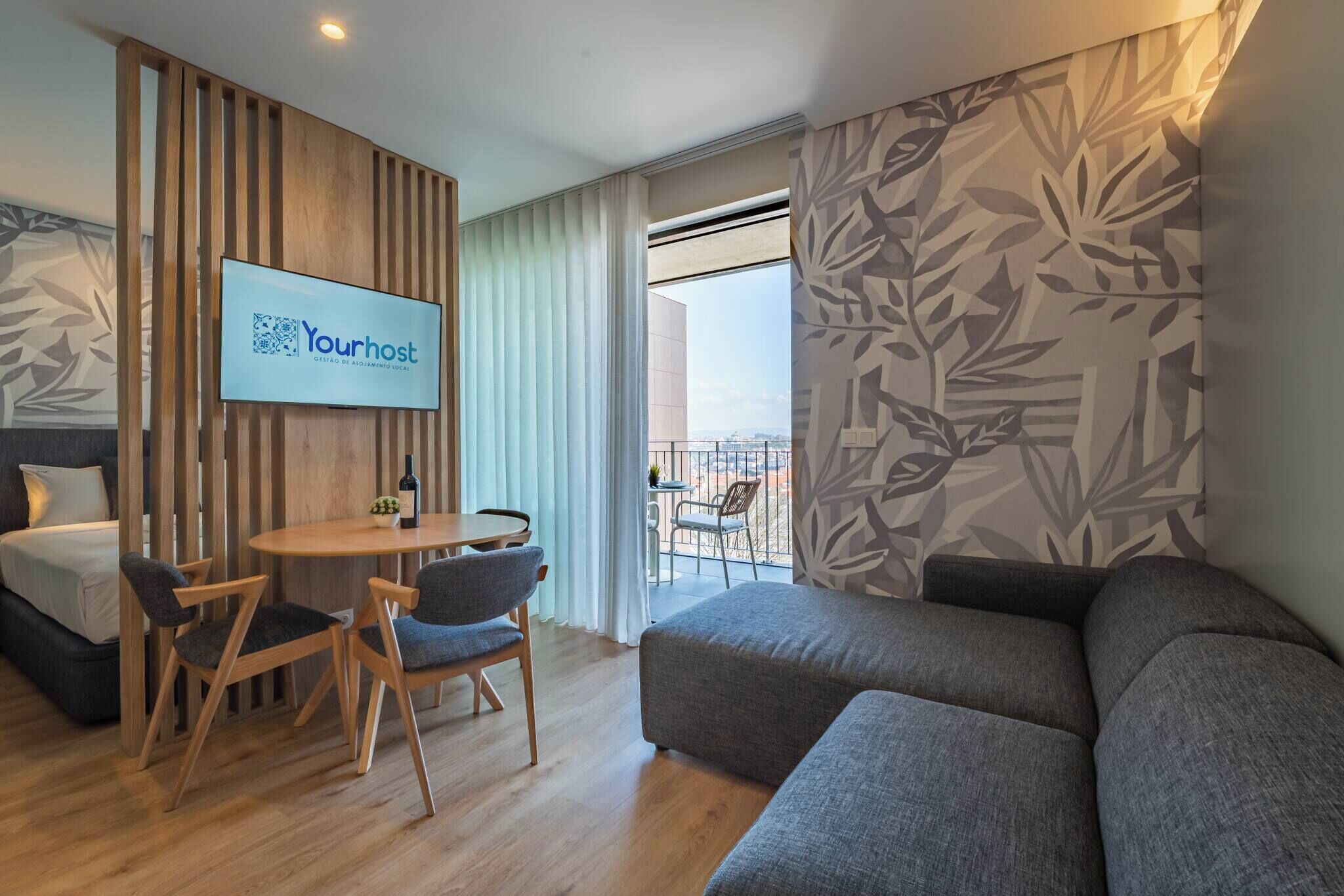A modern hotel room with a wooden partition, a flat-screen TV, a dining area with a table and chairs, a grey sofa, and a balcony with a view.