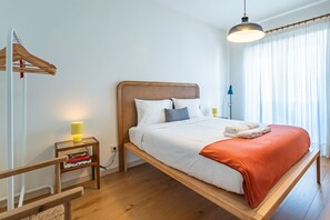 Un lit simple avec une tête de lit en bois, une table de chevet avec une lampe et une fenêtre ornée de rideaux légers.