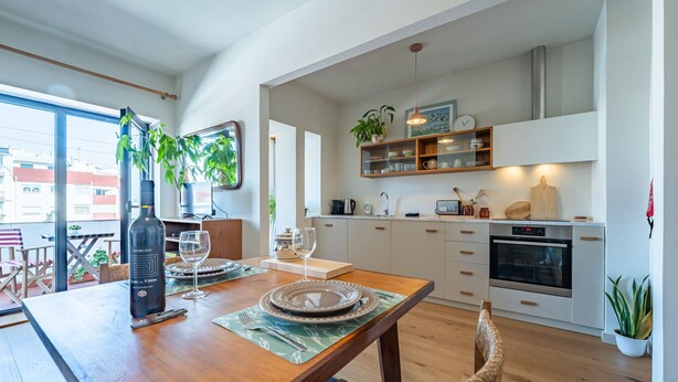 A modern kitchen with white cabinets, a dining area with a wooden table, and a view of the city through large windows. - Charming retreat near Boavista with elevator (Oporto)