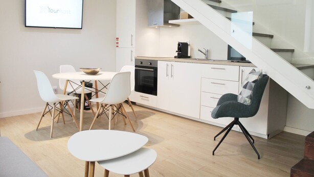 A modern kitchen with white cabinets, a black oven, and a coffee machine. There is a dining area with a white table and chairs, and a staircase leading to an upper level. - Typical Quarter Nest (Oporto)