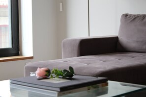 Ein modernes Wohnzimmer mit einem grauen Sofa, einem Glastisch und einem Fenster mit Blick auf Gebäude.