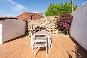 Un patio al aire libre con una mesa, sillas, un paraguas grande y una pared de piedra adornada con plantas en flor.