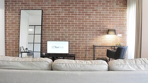 A living room with a brick wall, a large mirror, a flat-screen TV, a modern floor lamp, and a sofa. - Arrábida Luxury Studio (Vila Nova de Gaia)
