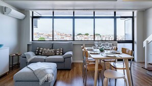 Un espacio habitable moderno con sofá, mesa de comedor y sillas, ventanas amplias con vista a la ciudad, y una pequeña mesita auxiliar.