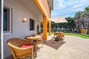 A covered outdoor seating area with wicker furniture and a table, a large umbrella, and a view of a lush garden. - Wake up with the Sea Breeze- House with pool AP2 (Lavra)