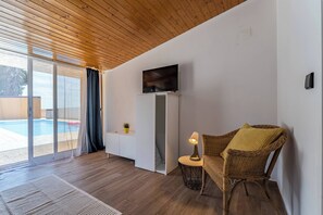 A room with a wooden ceiling, a television on a white cabinet, a wicker chair with a cushion, a small table with a lamp, and a sliding glass door leading to a pool area. - Wake up with the Sea Breeze- House with pool AP2 (Lavra)