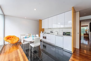 Cocina moderna con armarios blancos, zona de comedor con mesa y sillas blancas, y una silla de color naranja brillante.