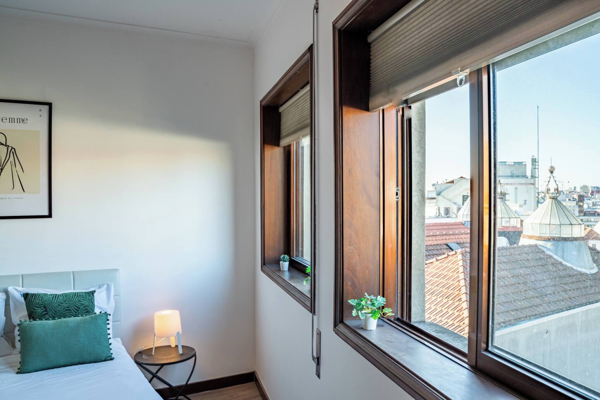 Habitación de hotel con cama, lámpara de noche, una mesita con lámpara, ventana con vista a la ciudad y un cuadro en la pared.