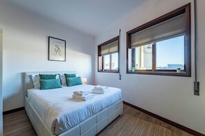 Ein Schlafzimmer mit Bett, Kissen, einem gerahmten Bild, einem Fenster mit Jalousien und Blick auf Gebäude.