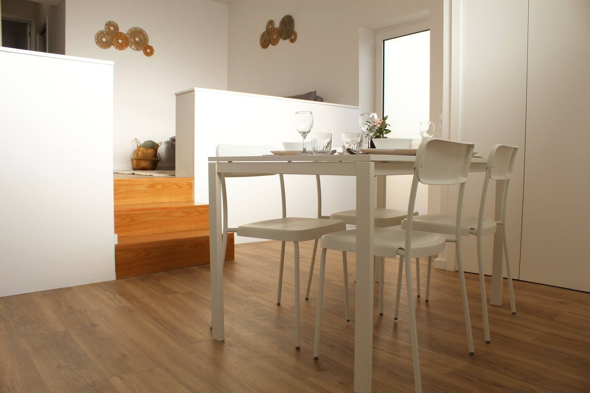 A dining area with a table and chairs, a wooden staircase, and a kitchen in the background.