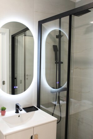 A modern bathroom with a glass-enclosed shower, a white sink with a black faucet, and a mirror with a circular light. - Valbom Hideaway Light, Style and Peace (Valbom)