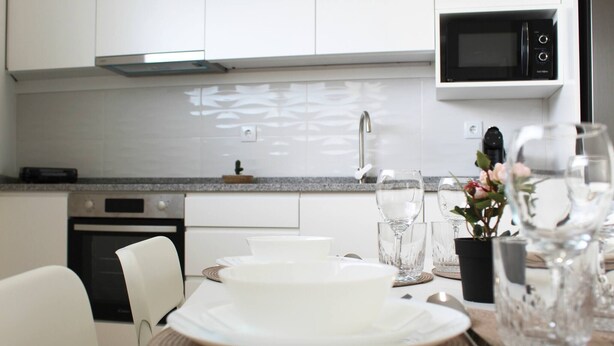 A modern kitchen with white cabinets, a microwave, an oven, and a dining area set with white plates, glasses, and a vase with flowers. - Valbom Hideaway Light, Style and Peace (Valbom)