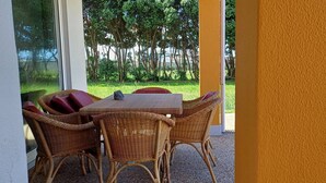 Een set rieten tafel en stoelen in een overdekt buitengebied, met uitzicht op bomen en een gebouw op de achtergrond.