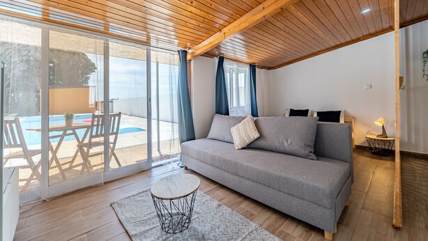 Een moderne woonkamer met een houten plafond, een grijze bank, een ronde salontafel en uitzicht op een zwembad door grote glazen deuren.