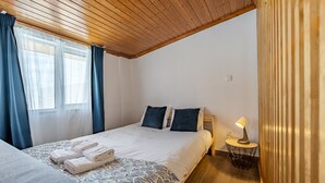 Een slaapkamer met een houten plafond, een bed met witte lakens, twee kussens, een nachtkastje met een lamp en een raam met gordijnen.