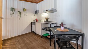 Een kleine keuken met een houten plafond, witte muren, een magnetron, een spoelbak, een kleine koelkast, een eettafel met stoelen en hangende planten.