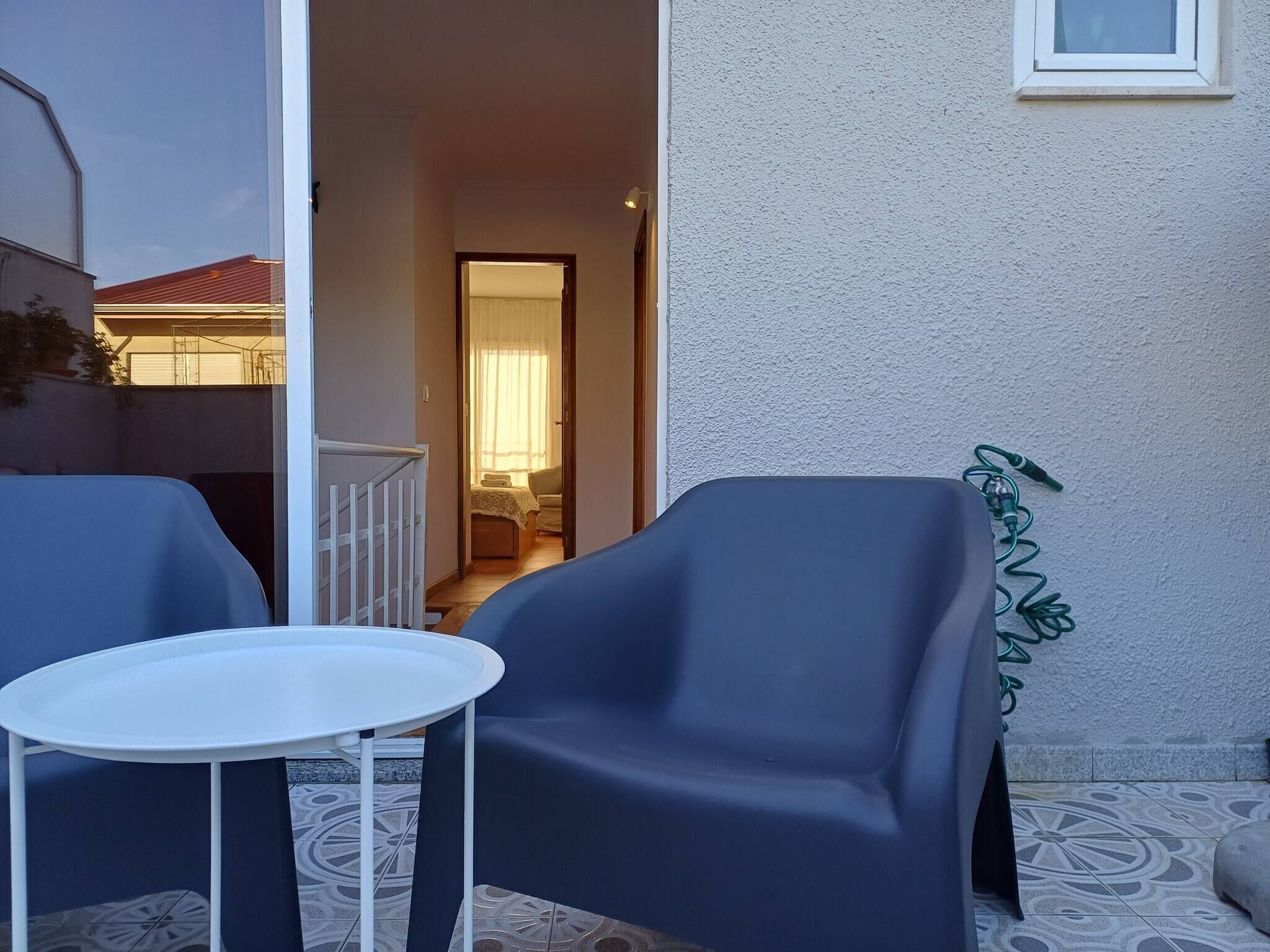 Un patio con una silla azul y una pequeña mesa blanca, una puerta que da a una habitación interior, y una lámpara fijada a la pared.