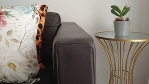 Un sofá de cuero oscuro con un cojín floral, una pequeña planta en maceta sobre una mesita de alambre dorado y una pared lisa.