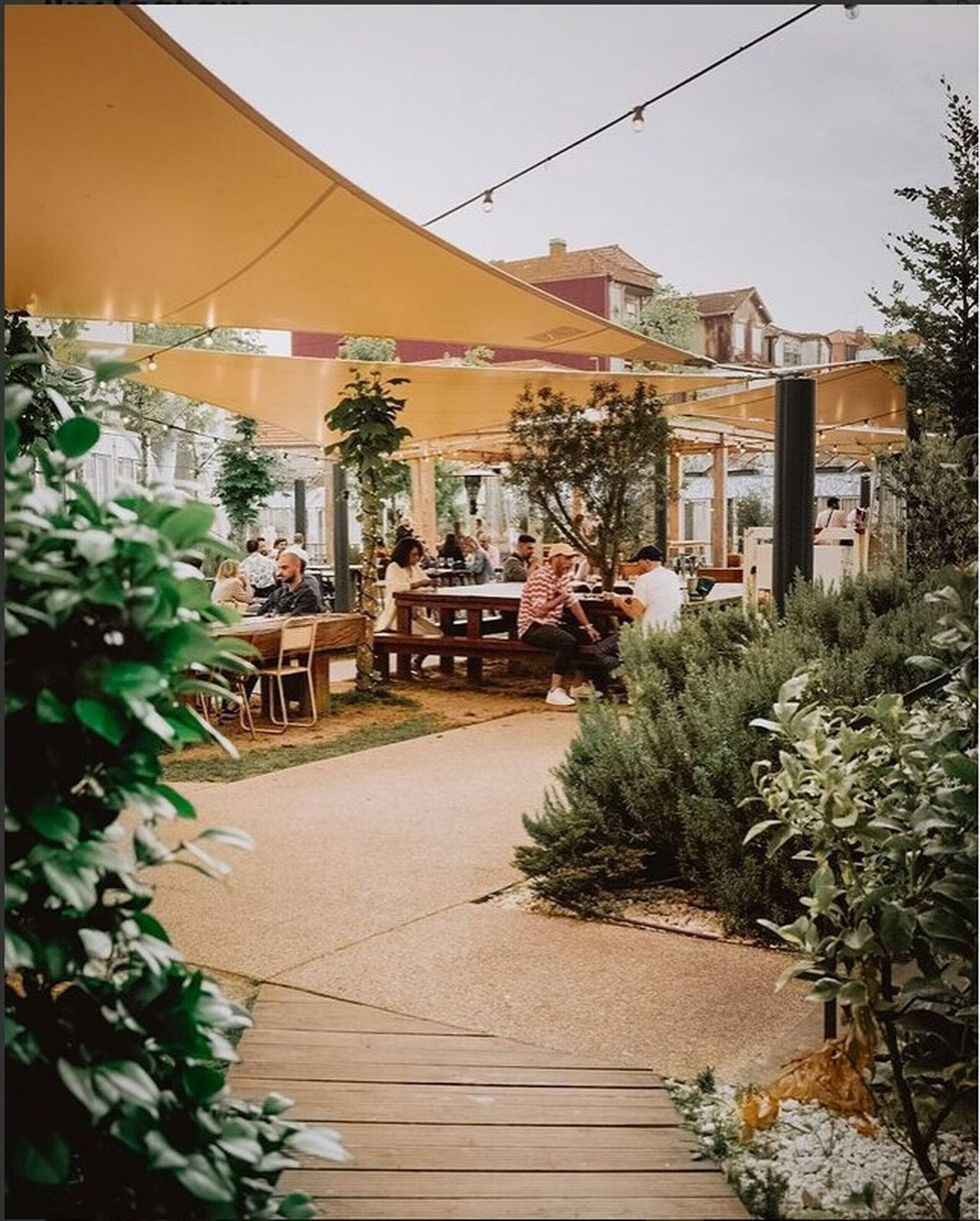 An outdoor seating area with tables and chairs under a large canopy, surrounded by greenery.