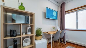 Une pièce avec une bibliothèque en bois, un petit réfrigérateur blanc, un bureau avec une chaise, un téléviseur fixé au mur et une fenêtre avec des rideaux.