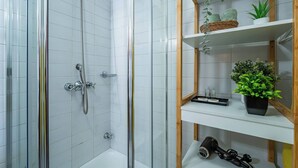Une douche avec une porte en verre, un mur recouvert de carreaux blancs et une étagère avec des plantes et un sèche-cheveux.