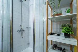 Eine Dusche mit Glastür, eine weiße Fliesenwand und ein Ablagebereich mit Pflanzen und einem Fön.