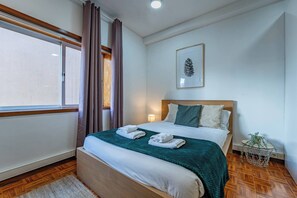 Ein Hotelzimmer mit Bett, einem Vorhangfenster, einem gerahmten Bild an der Wand und einem kleinen Nachttisch mit Pflanze.