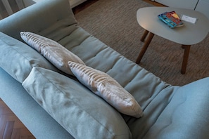 Ein hellgraues Sofa mit einem großen, strukturierten Kissen und einem kleinen Tisch mit einem Buch.