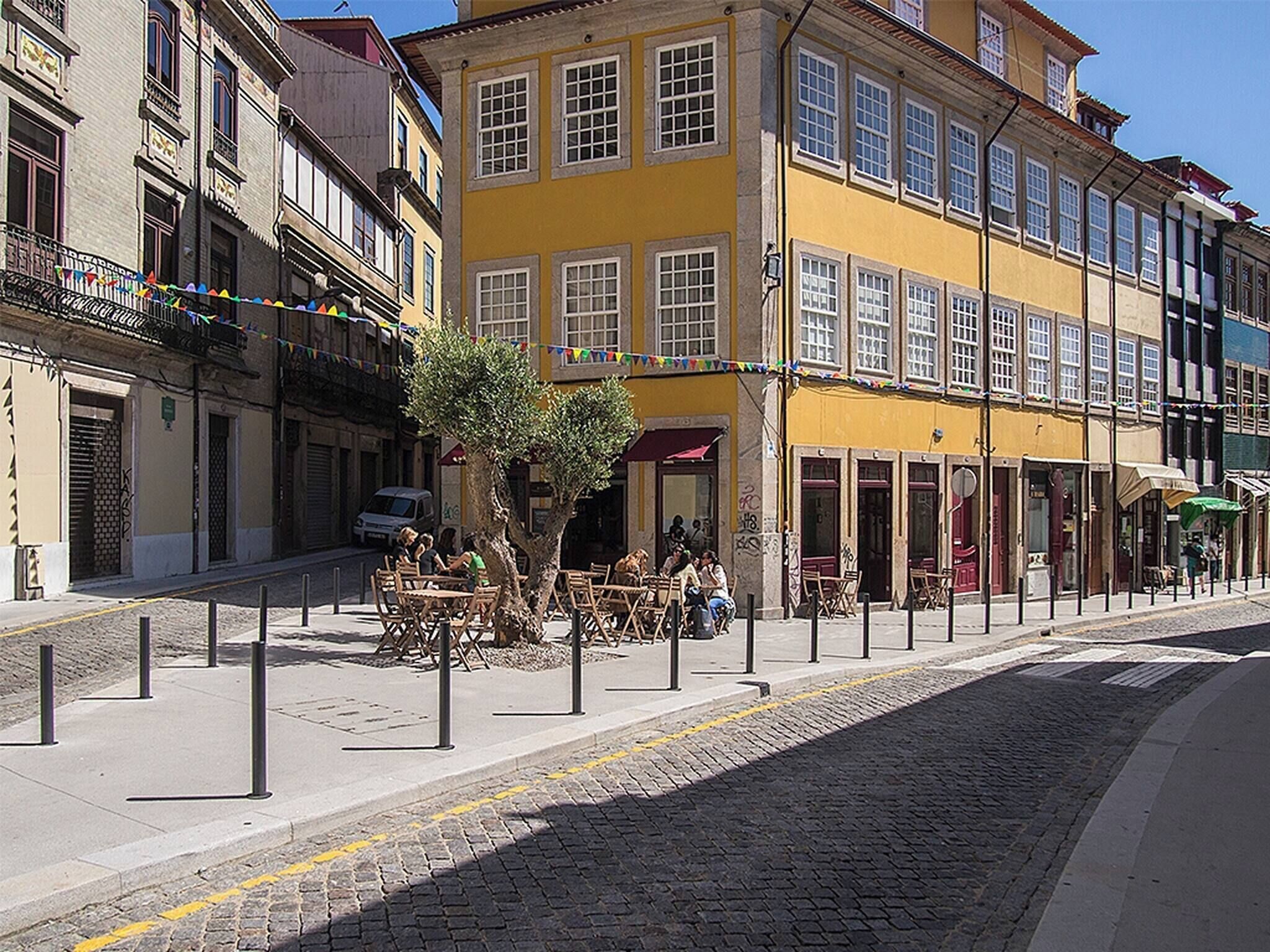 Une rue bordée d’immeubles, avec un arbre et des tables installées à l’extérieur.