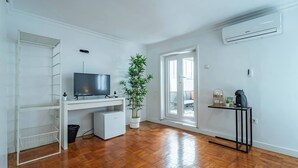 Una habitación con piso de madera, un televisor sobre un escritorio, una planta en maceta, una mesita con cafetera y un equipo de aire acondicionado.
