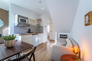 Eine moderne Küche mit weißen Schränken, einer Mikrowelle und einer Kaffeemaschine. Es gibt eine Essecke mit einem Holztisch und Stühlen, sowie ein weißes Sofa mit einer Lampe.