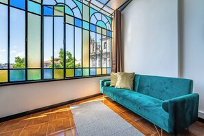 Ein Zimmer mit einem türkisfarbenen Sofa, einem großen Fenster mit Buntglas und Blick auf ein Gebäude.