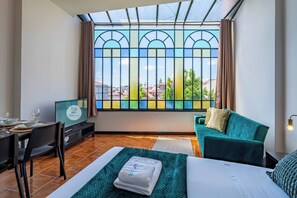 Ein Hotelzimmer mit großem Fenster, einem Bett, einem grünen Sofa, einem Fernseher und einer Essecke mit Stühlen und Tisch.