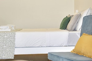 Een netjes opgemaakt bed met witte lakens, een grijze bank met een geel kussen, en een geborduurd plaid aan de linkerkant.