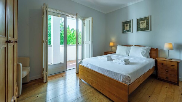 A bedroom with a wooden bed, a nightstand with a lamp, a chair, and a sliding glass door leading to an outdoor area. - Casa das Pintoras Garden and Pool (Vile)