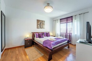 A bedroom with a wooden bed, a bedside table, a desk with a computer, a window with curtains, and a painting on the wall. - Cozy and Quiet Spacious Flat (Oliveira do Douro)