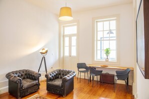 Ein Zimmer mit Holzboden, zwei Ledersesseln, einem Schreibtisch mit Stühlen, einer Stehlampe und einem Fenster mit Pflanze.