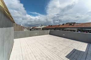 Eine Dachterrasse mit Fliesenboden, einer Betonwand und Blick auf Gebäude mit roten Ziegeldächern.