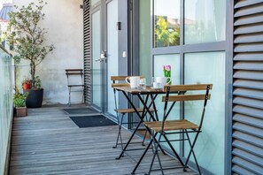 Una terrassa de fusta amb taula i cadires, plantes en testos i vista d'un edifici a través d'una porta de vidre.