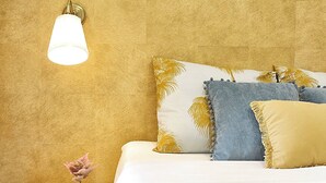 Una cama con colcha blanca y dorada, un almohadón azul y un almohadón amarillo con flecos. Hay una mesita de noche con un libro y una pequeña planta en maceta.