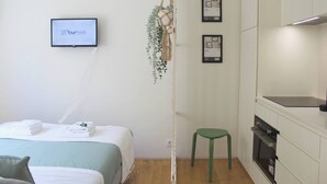Hotellihuone, jossa on sänky, vihreä jakkara, seinälle asennettu televisio, jossa näkyy sovellus nimeltä »Yourhost», sekä keittiöalue, jossa on kaappeja ja uuni.