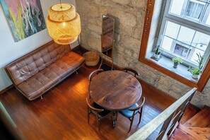 Ein Zimmer mit einem braunen Ledersofa, einem runden Holztisch mit Stühlen, einer hängenden Korbtasche, einem Spiegel und einem Fenster mit Pflanzen.