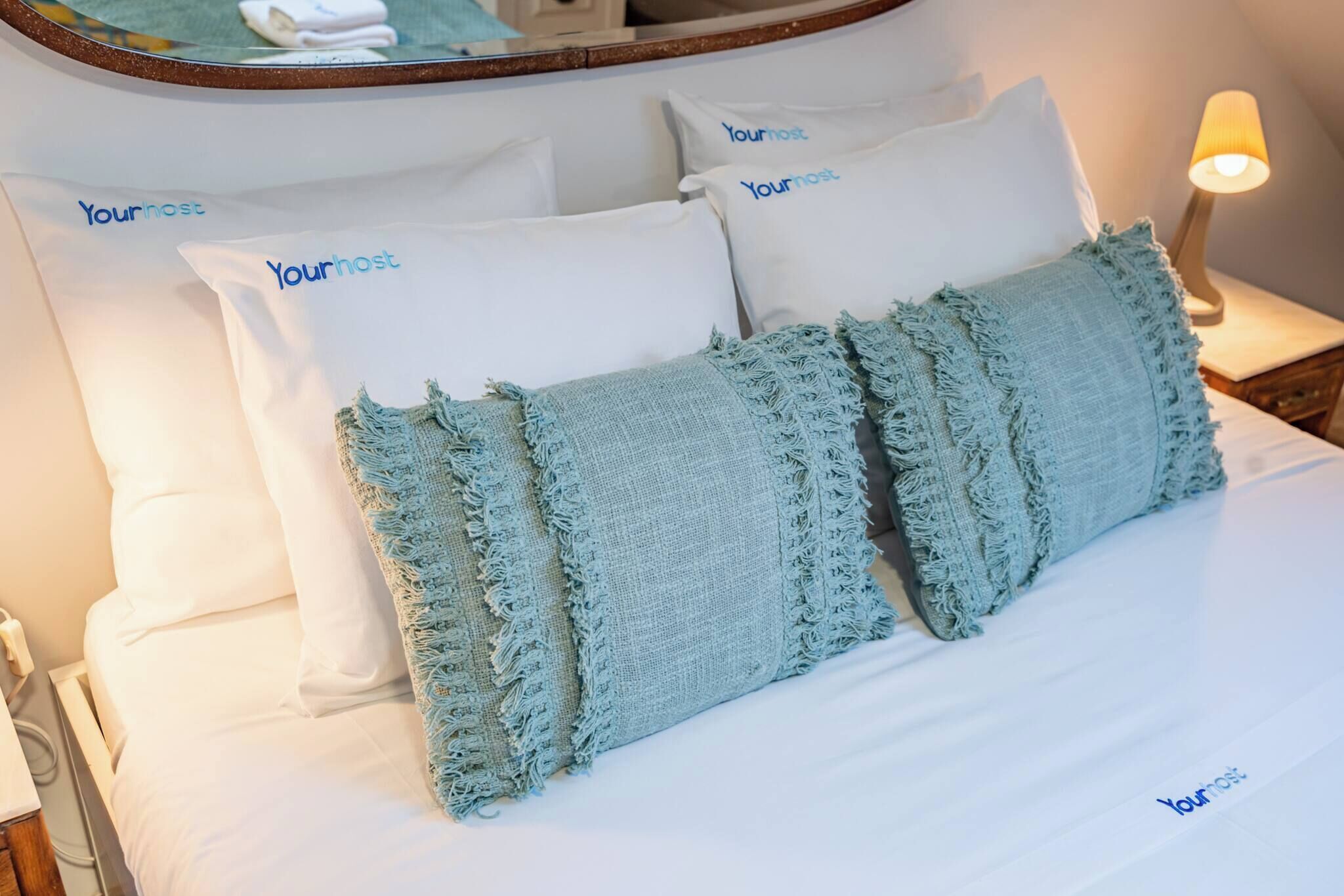 Una cama bien tendida con almohadas que dicen 'Yourhost', una manta texturizada azul claro y una lámpara de noche.