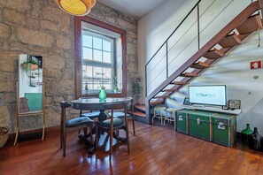 Ein gemütliches Zimmer mit Steinwand, Holzboden, einem runden Tisch mit Stühlen, einem Fenster, einer Treppe und einem Fernseher auf einem grünen Koffer.