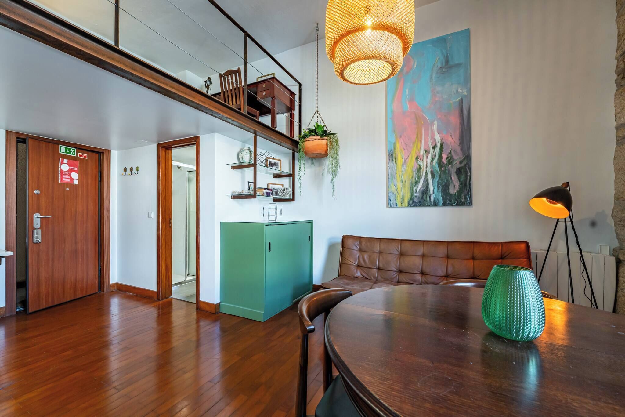 Une pièce spacieuse avec un plancher en bois, une table à manger avec des chaises, un canapé, une plante suspendue, un tableau au mur et un escalier menant à un niveau supérieur.
