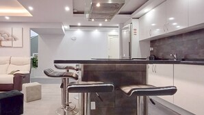 Ein modernes Wohnzimmer mit einer Couch, einem Couchtisch und einer Küchenzeile inklusive Barhocker und Kühlschrank.