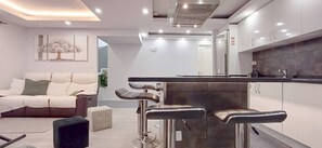 Ein modernes Wohnzimmer mit einer Couch, einem Couchtisch und einer Küchenzeile inklusive Barhocker und Kühlschrank.