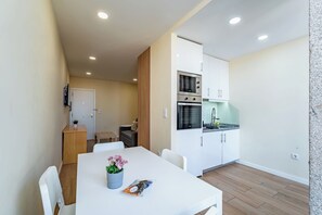 A compact kitchen with white cabinets, a microwave, and a stove. There is a dining area with a white table and chairs, and a small wooden side table with a plant. - Close to Everything Your Oporto Gray PRM 2T (Porto)