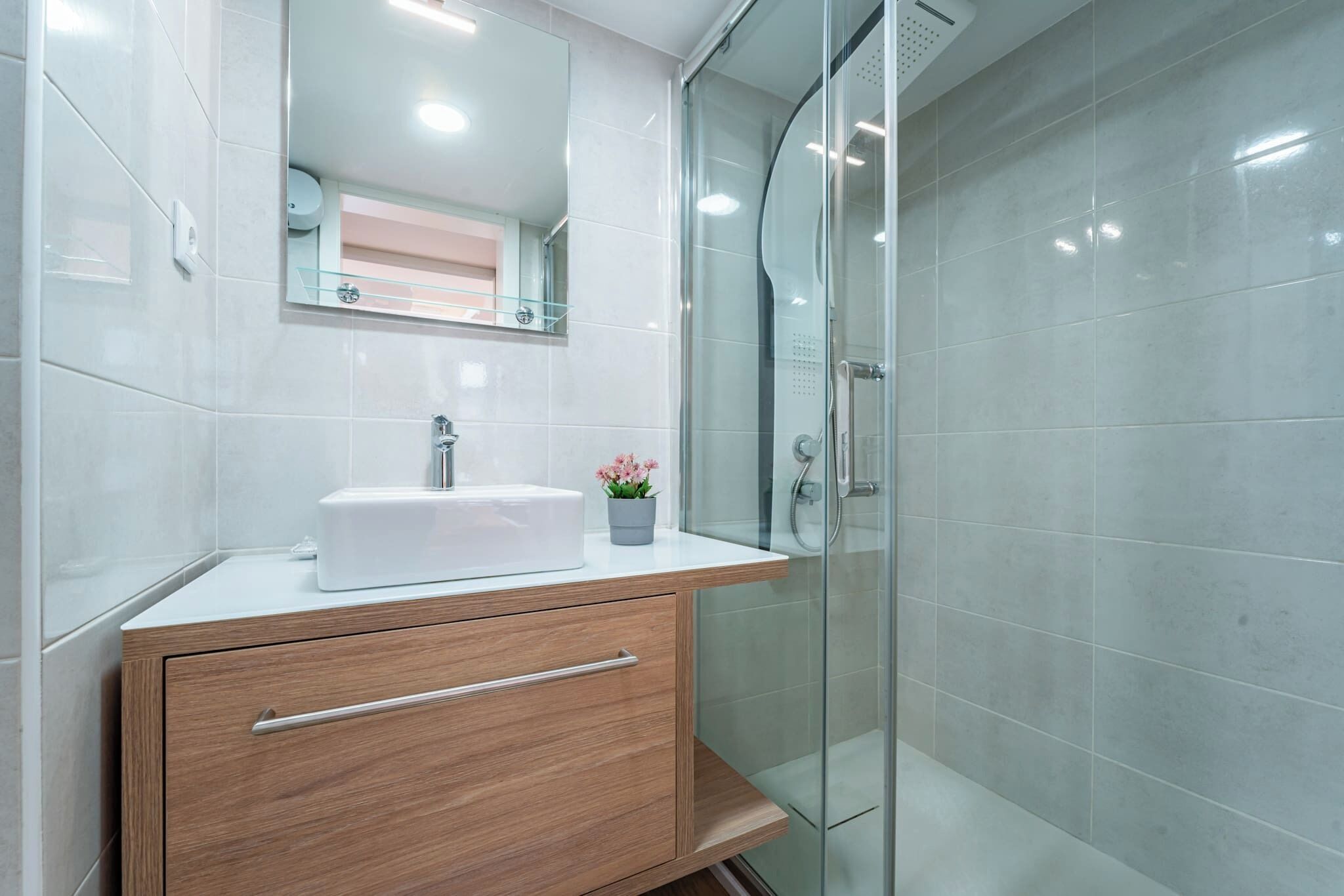 Un baño moderno con lavamanos blanco, tocador de madera y cabina de ducha de vidrio.