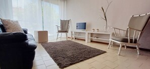 Een woonkamer met een bank, twee fauteuils, een televisie, een witte console tafel en een groot raam.